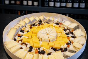 Sélection de fromages artisanaux et fruits secs sur un plateau en bois circulaire devant une cave à vin.