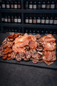 Plateau de charcuterie fine avec jambon cru, saucisson et chorizo sur ardoise devant une cave à vin.