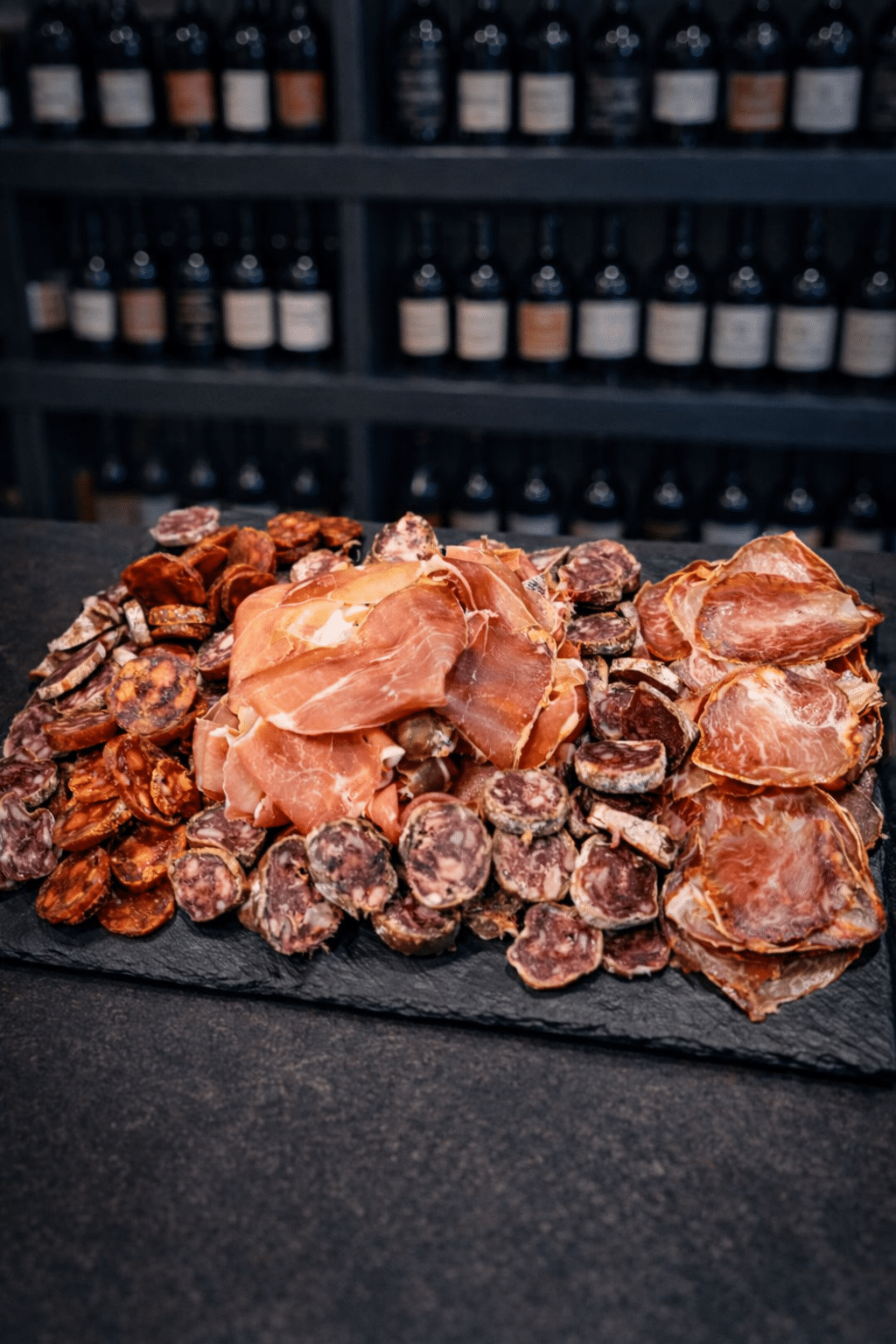 Plateau de charcuterie fine avec jambon cru, saucisson et chorizo sur ardoise devant une cave à vin.