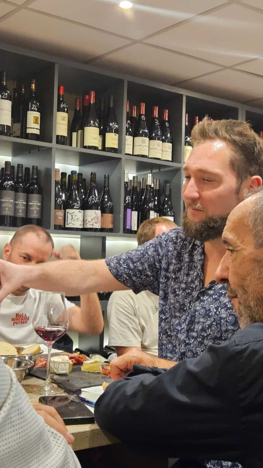 Soirée Dégustation de Vin Rouge et Fromages au Bar Dégustation conviviale de vin rouge et fromage au bar. Homme en t-shirt 'Not a morning person' devant la cave.
