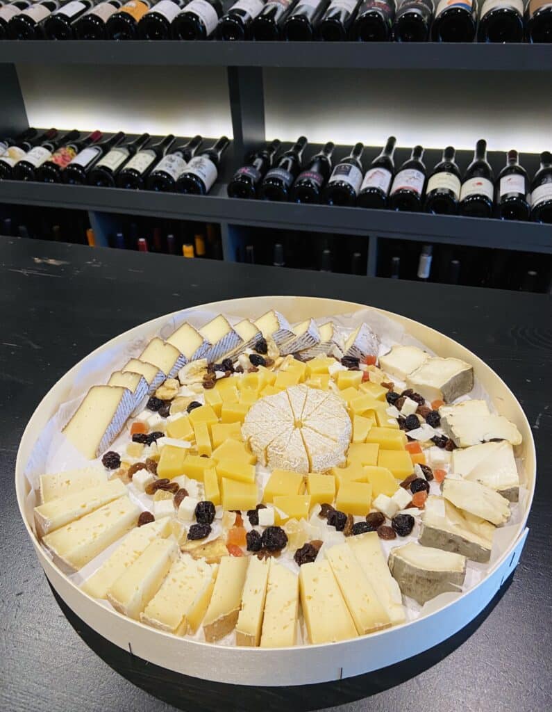 Plateau de Fromages Assortis et Sélection de Vins Fins Assortiment gourmand de fromages et fruits secs présenté sur un plateau rond devant une cave à vin.